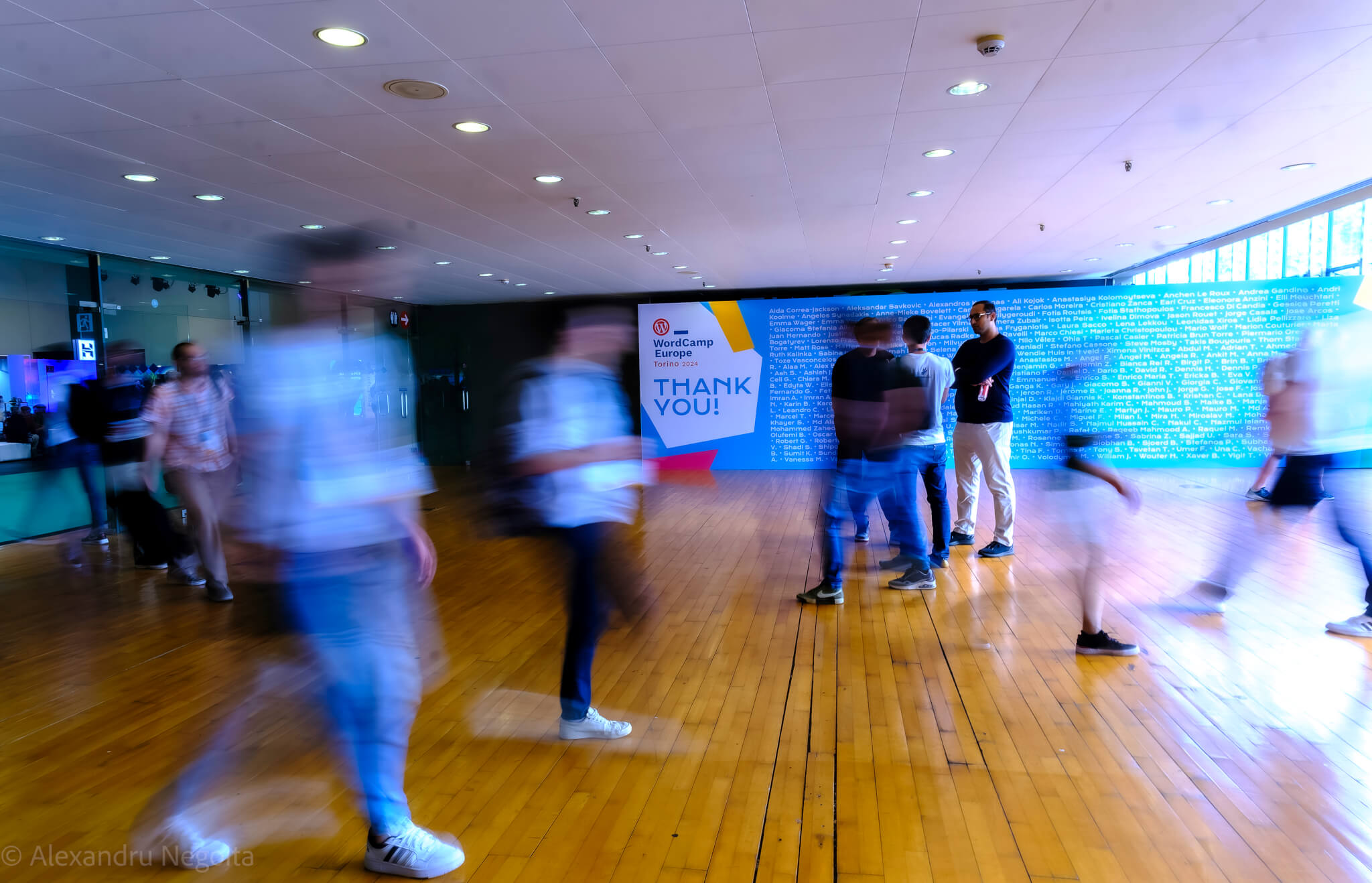 A picture I've taken in 2024, at WordCamp Europe, Torino (2024) with people crossing in front of the camera, with long exposure. On the background it is seen the wall with the people that helped creating and managing the event (organizers, volunteers including me, speakers and others).